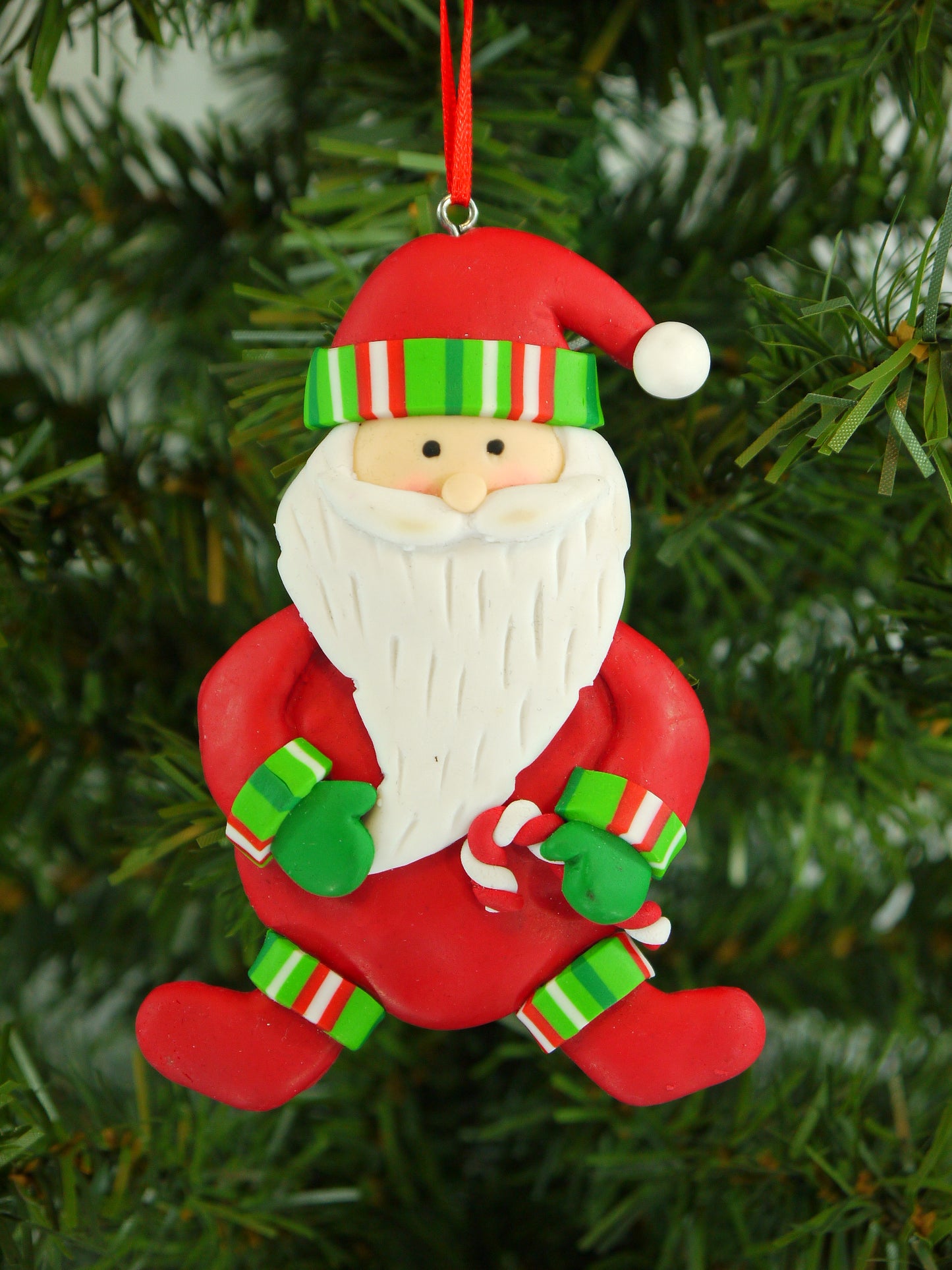 Detailed handmade polyclay Santa ornament holding a candy cane, hung on a green Christmas tree.