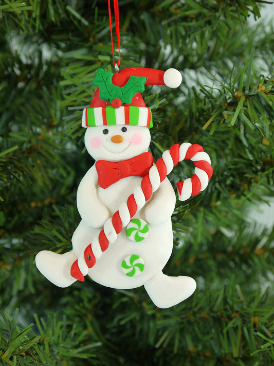 Detailed shot of a handmade polyclay snowman ornament with a candy cane, hanging on a green Christmas tree.