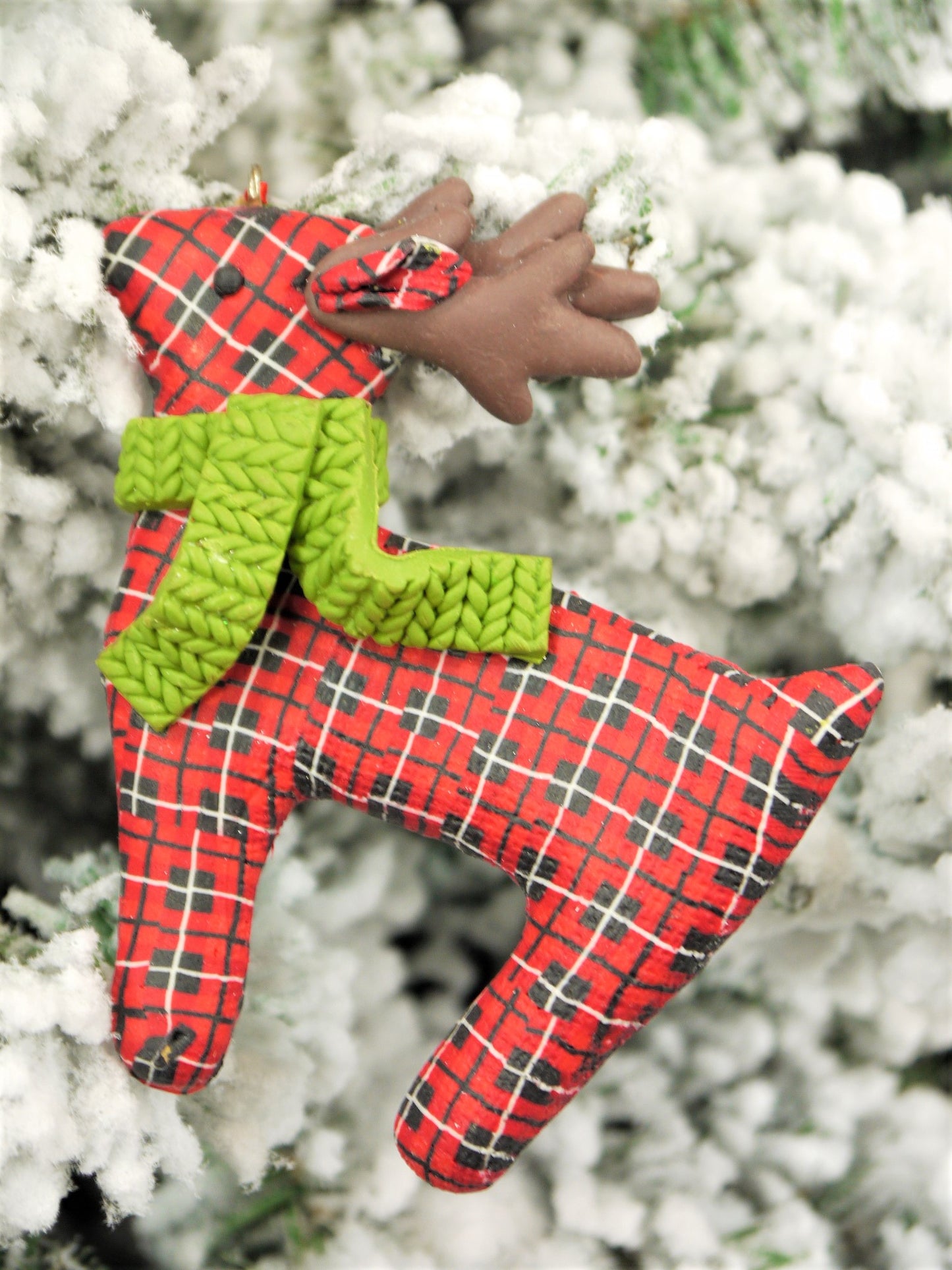 Handmade red plaid polyclay reindeer with a green knit scarf, hanging on a snow-covered Christmas tree.